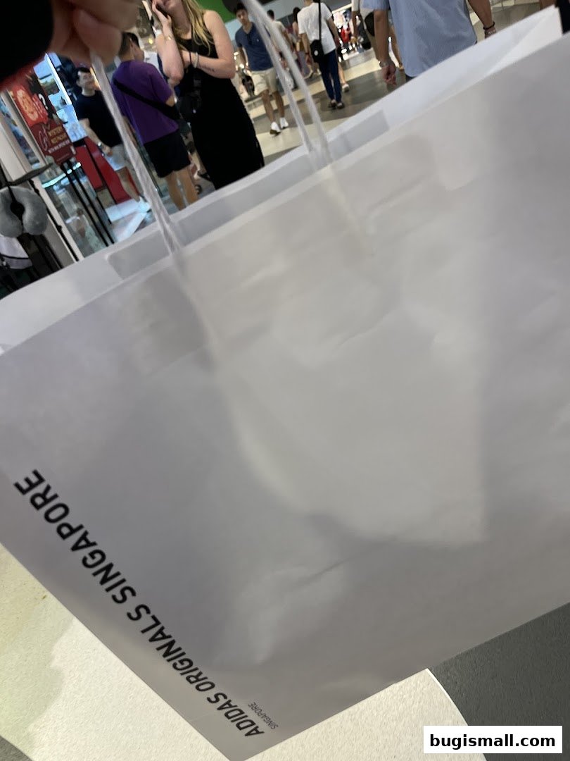 A white shopping bag with the Adidas Originals Singapore logo, held in a mall.