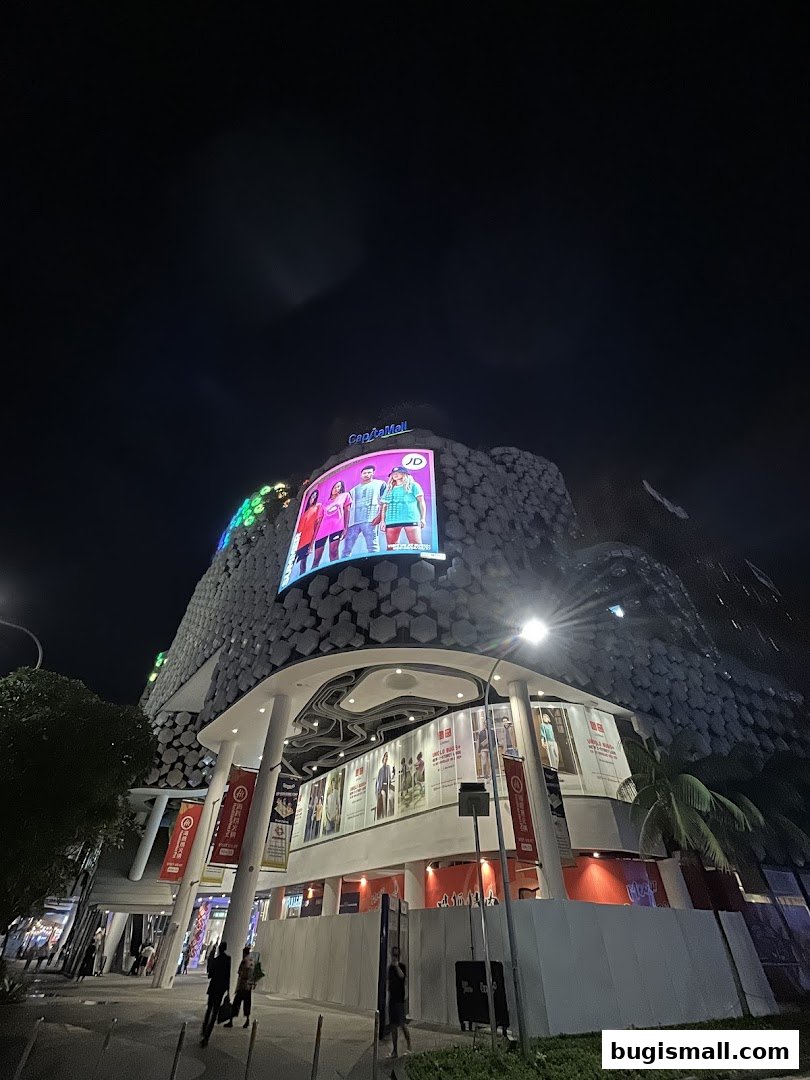 A nighttime view of the CapitaMall Bugis building with illuminated advertisements and storefronts.