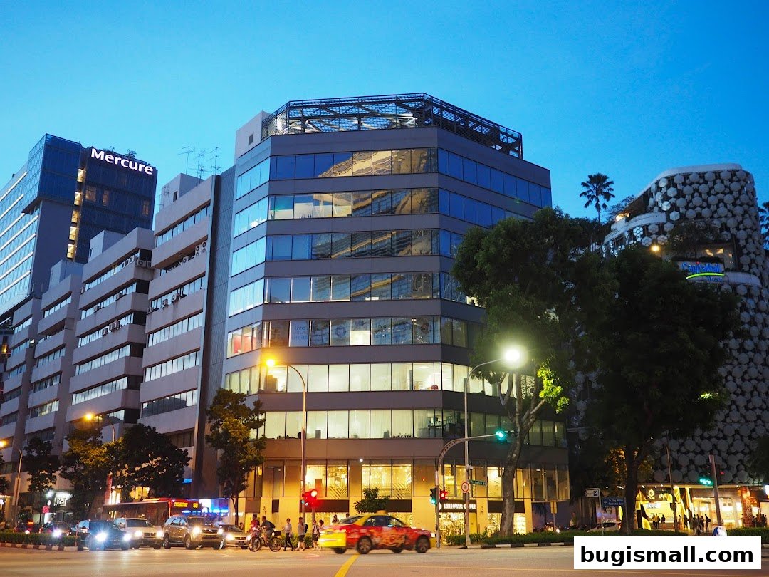 A street view of modern buildings at dusk, with traffic and illuminated windows.