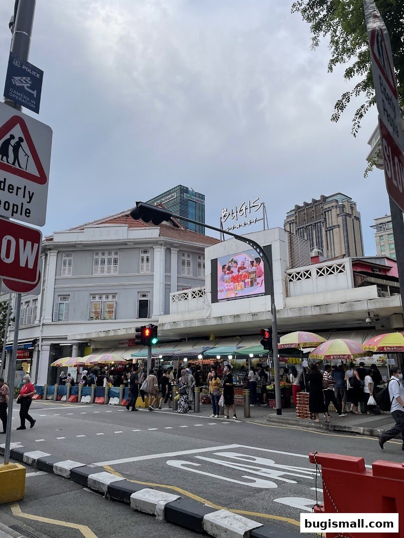 A street scene with a bustling market under awnings, with buildings and a large screen in the background.