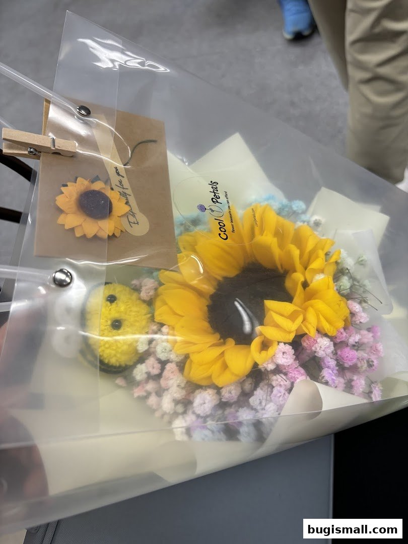 A bouquet featuring a large sunflower, baby's breath, and a small bee charm, presented in a clear bag.