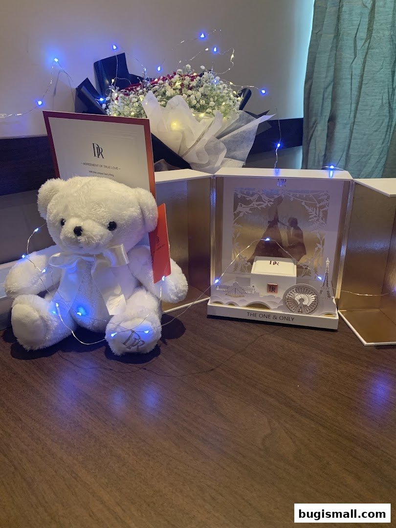A white teddy bear sits next to a bouquet of flowers and a gift box with fairy lights.