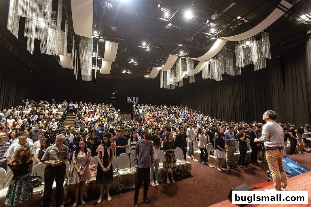 A large audience is seated in a hall with decorative ceiling elements, listening to a speaker.
