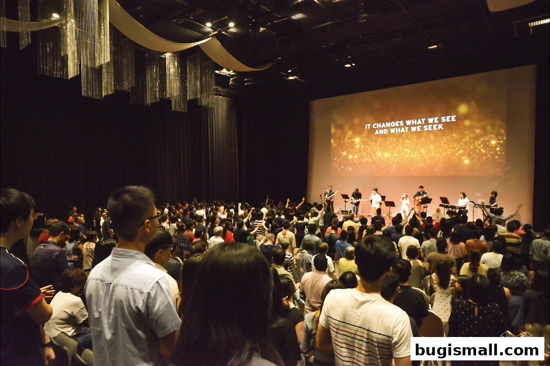 A large congregation watches a band perform on stage with lyrics displayed on a screen.