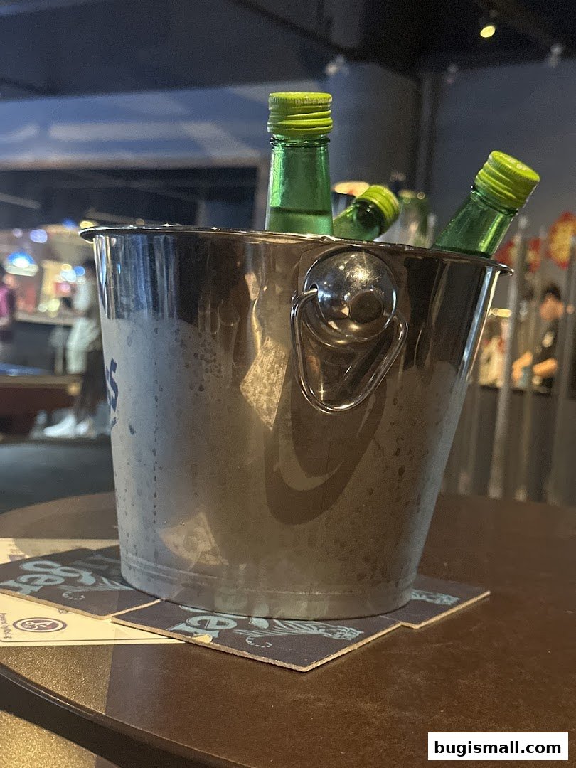 A metal ice bucket filled with green bottled drinks sits on a table with coasters.