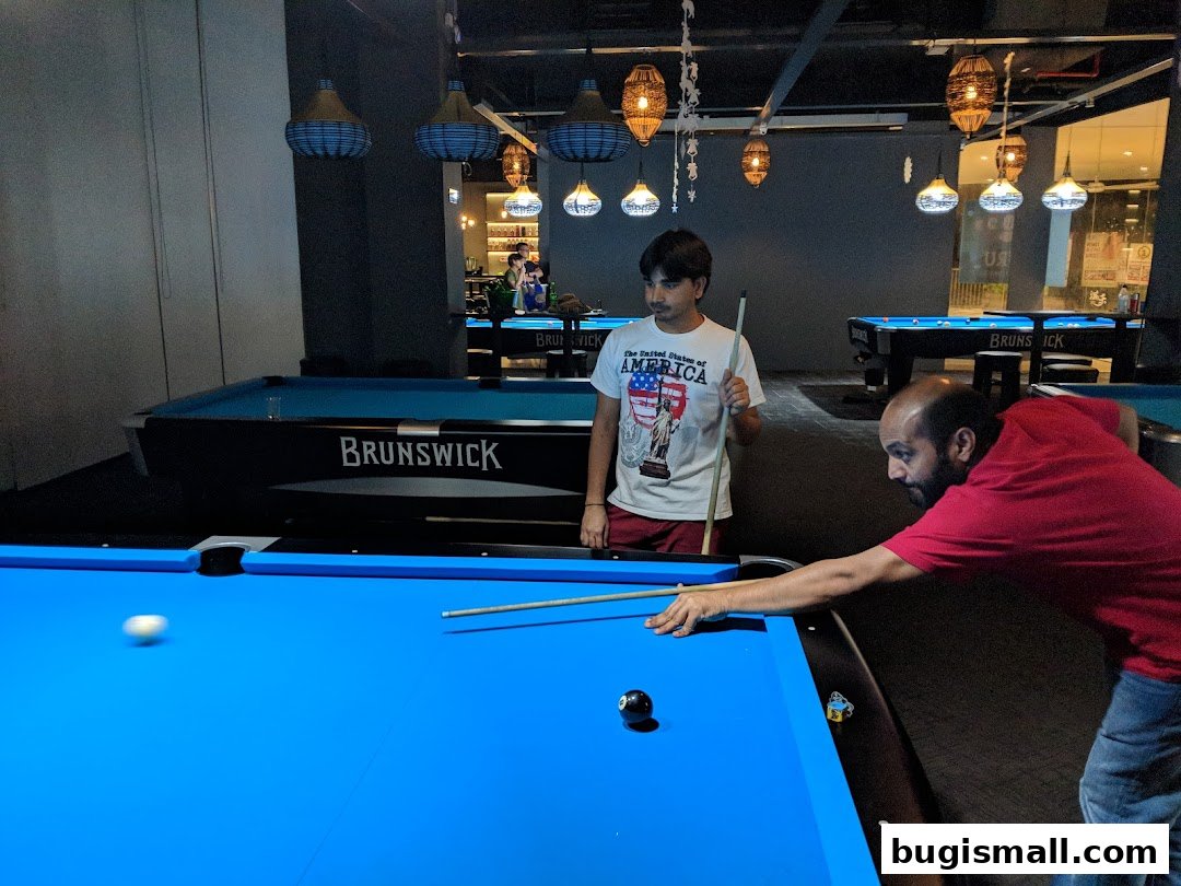 Two men playing billiards on a blue pool table in a dimly lit room with multiple tables.