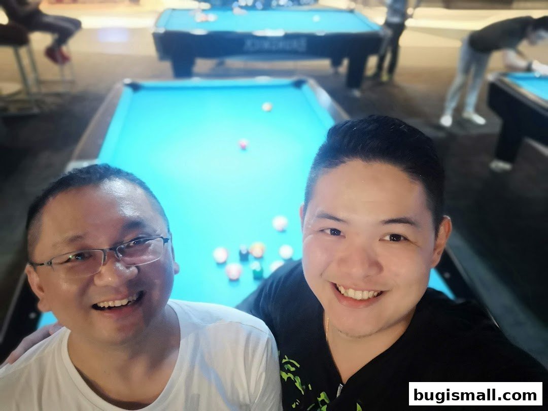 Two smiling men pose for a selfie in front of pool tables with other patrons playing.