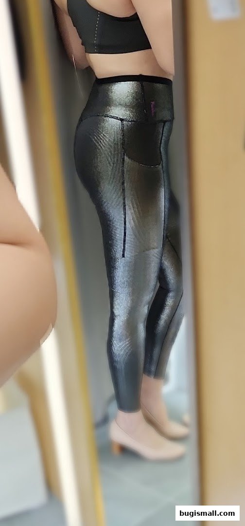 A person wearing shiny black leggings and a black sports bra is reflected in a mirror.