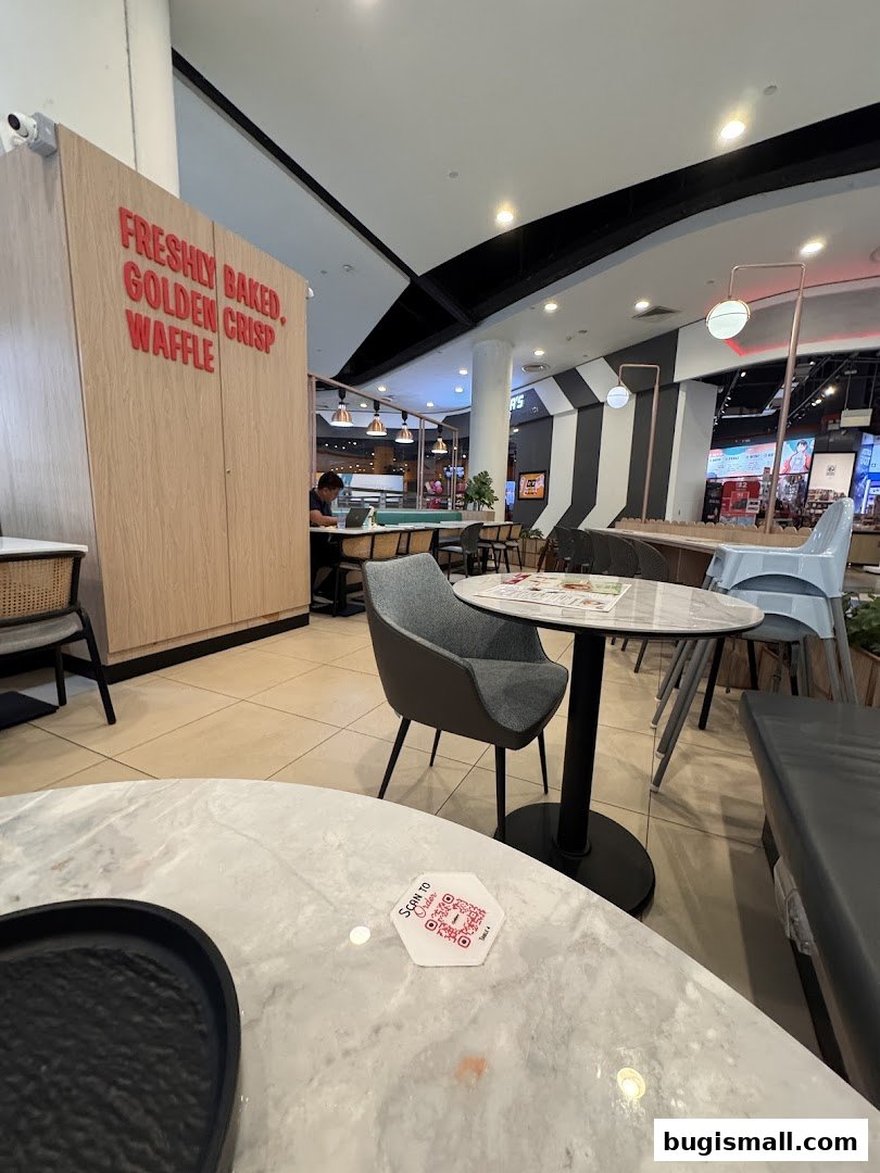 Interior of Geláre Bugis with tables, chairs, and a sign saying 'Freshly Baked, Golden Crisp Waffle'.