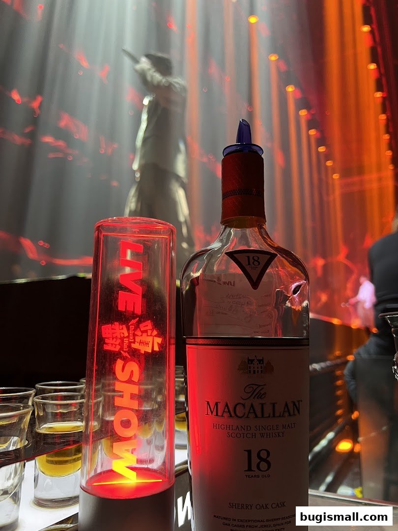 A bottle of Macallan 18 Year Old Scotch whisky and a 'Live Show' light stand are on a bar with a performer in the background.