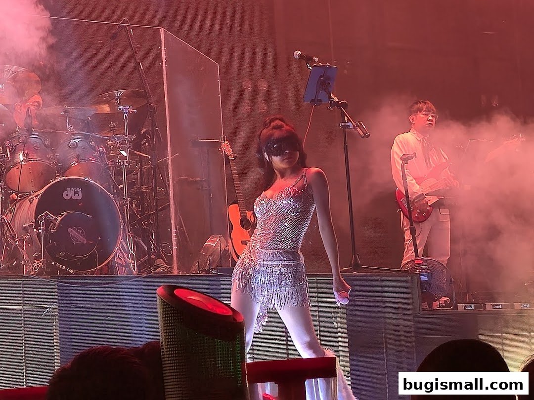 A female performer in a sparkly outfit sings on stage with a band.