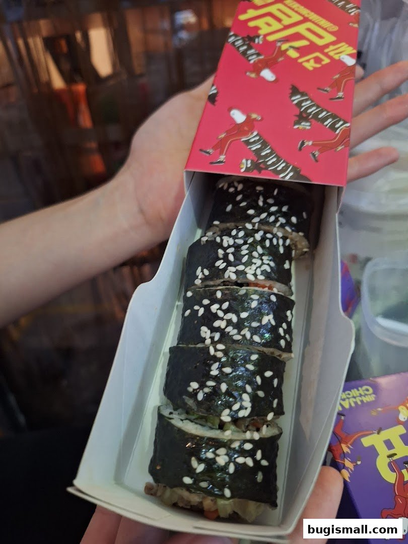 A hand holds a box of Korean kimbap rolls topped with sesame seeds.