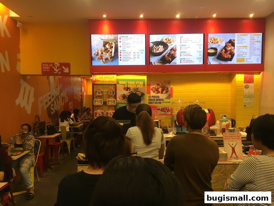 Customers queue at JINJJA Chicken Bugis Plus, with digital menus displaying food items.