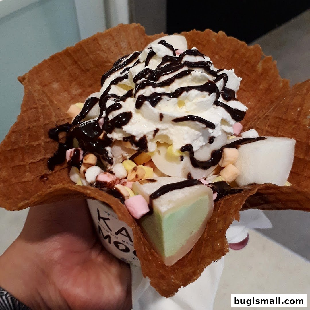 A hand holds a waffle cone filled with ice cream, whipped cream, chocolate sauce, marshmallows, and mochi.