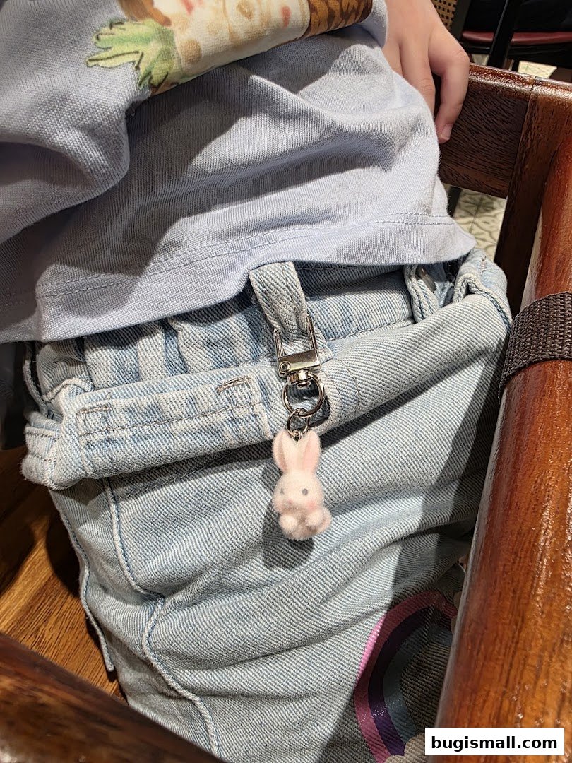 A close-up of a child's denim overalls with a fluffy pink bunny keychain attached.