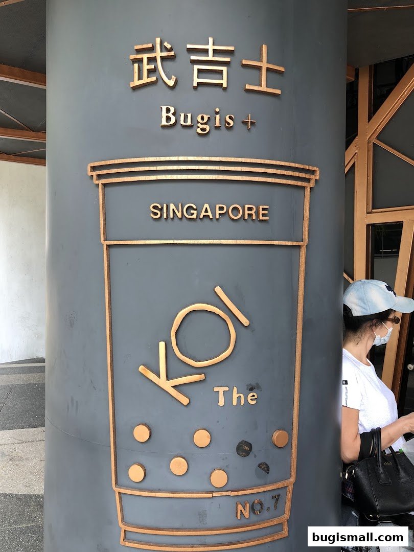 A pillar with the KOI Thé logo and "Bugis + Singapore" text, a woman stands nearby.