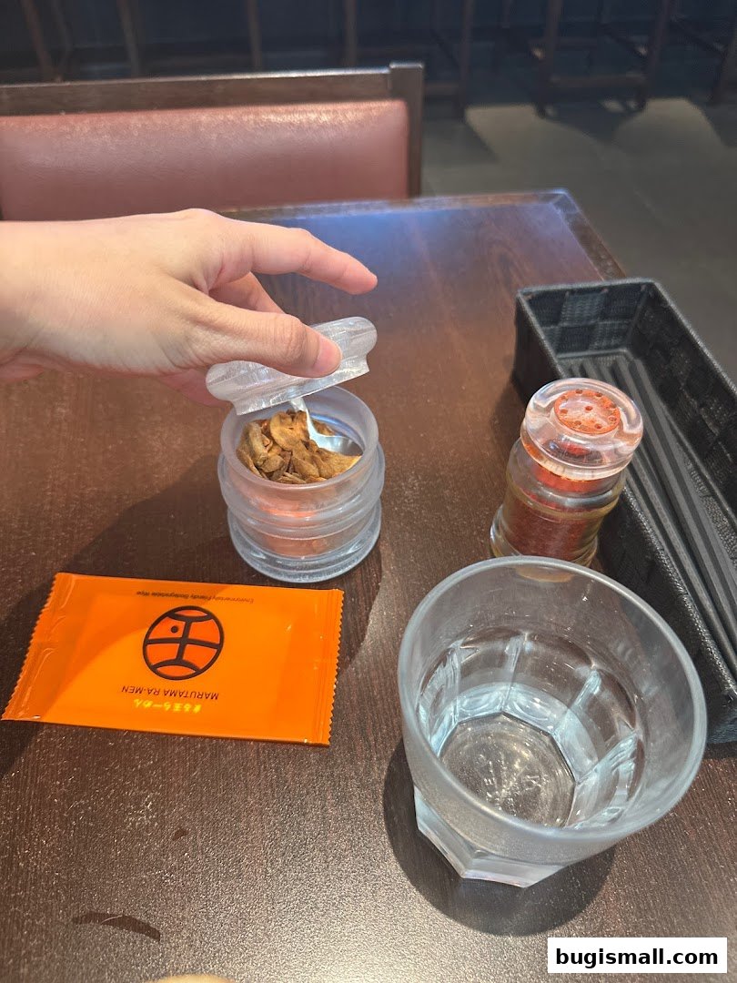 A hand opens a jar of fried garlic on a table at Marutama Ramen Bugis.