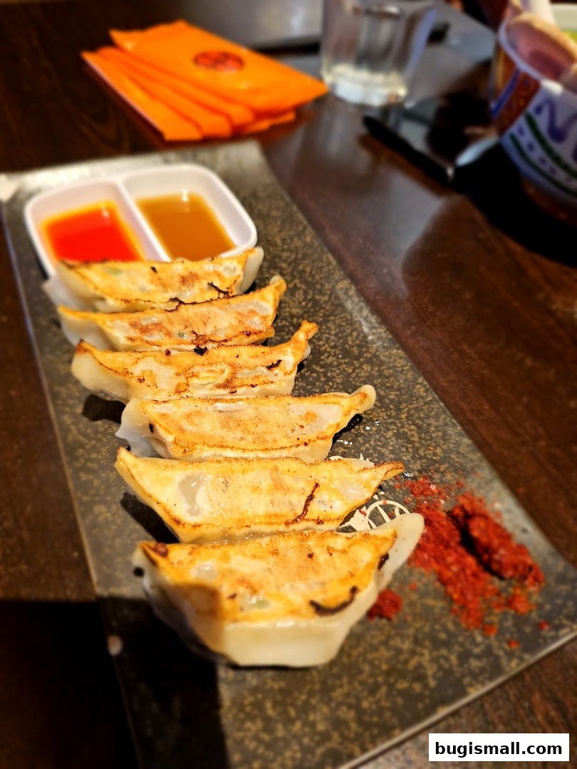 A plate of golden-brown pan-fried gyoza dumplings with dipping sauces and chili powder.