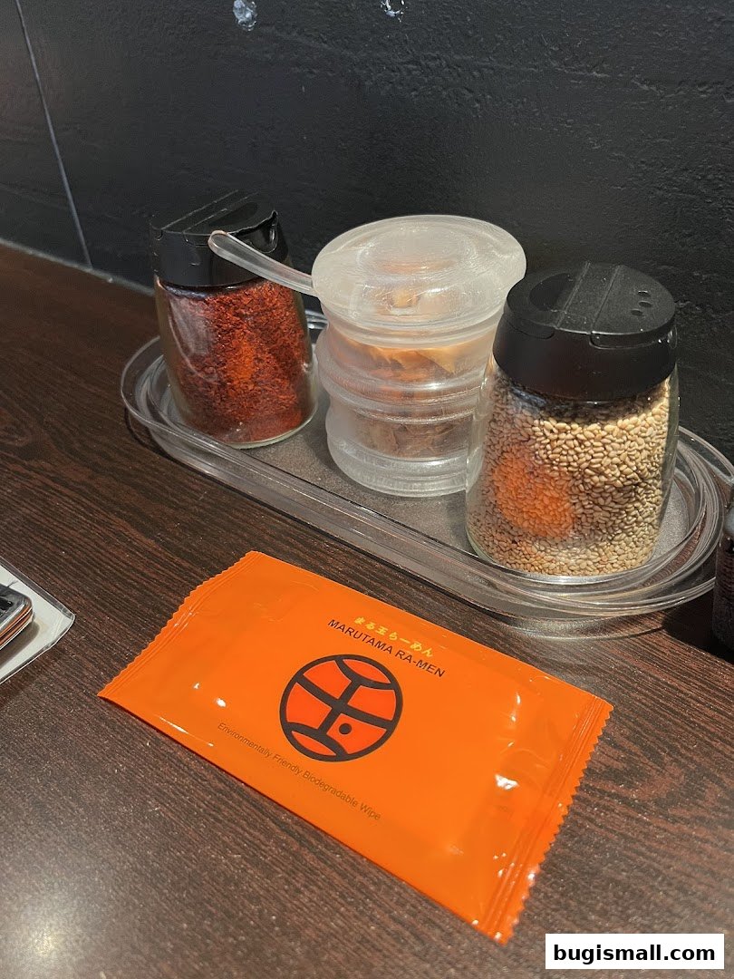 A table setting at Marutama Ramen Bugis with chili flakes, sesame seeds, and a wet wipe.