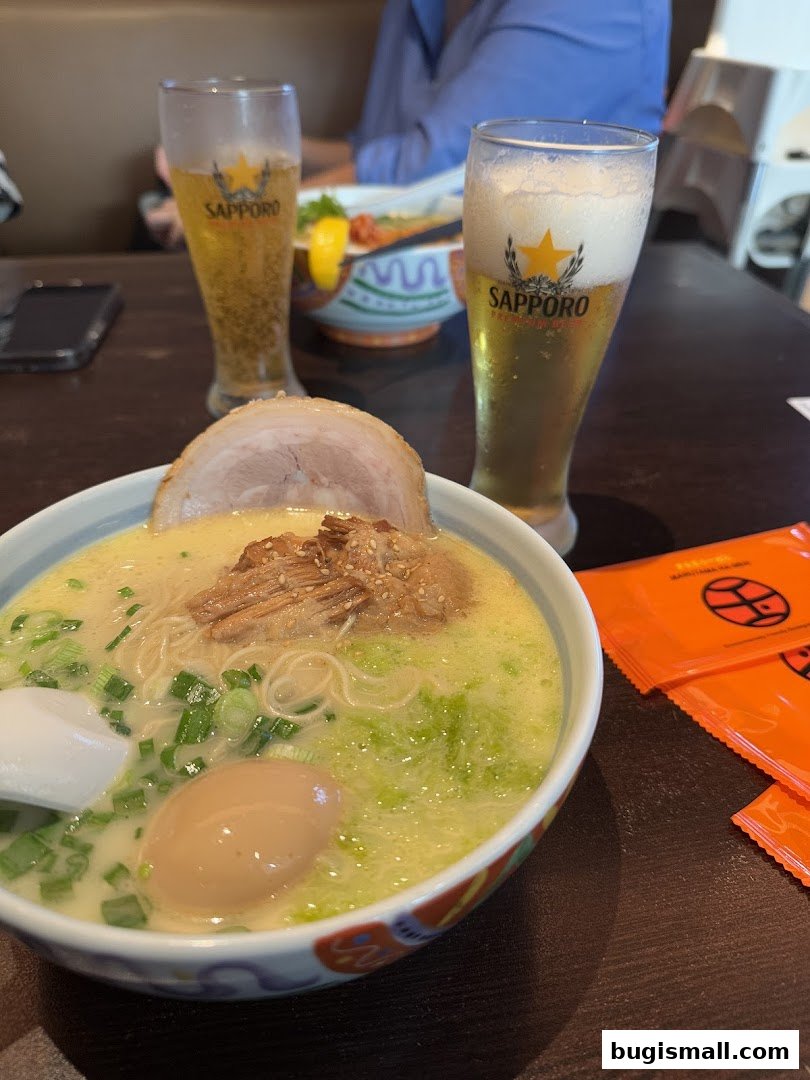 A bowl of creamy ramen with pork, egg, and green onions, served with two glasses of Sapporo beer.