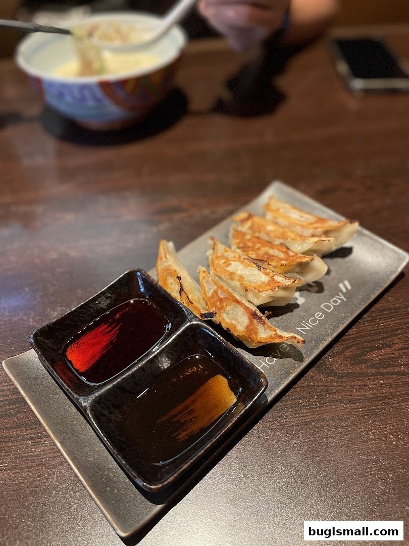 A plate of golden-brown gyoza with two dipping sauces and a bowl of ramen in the background.