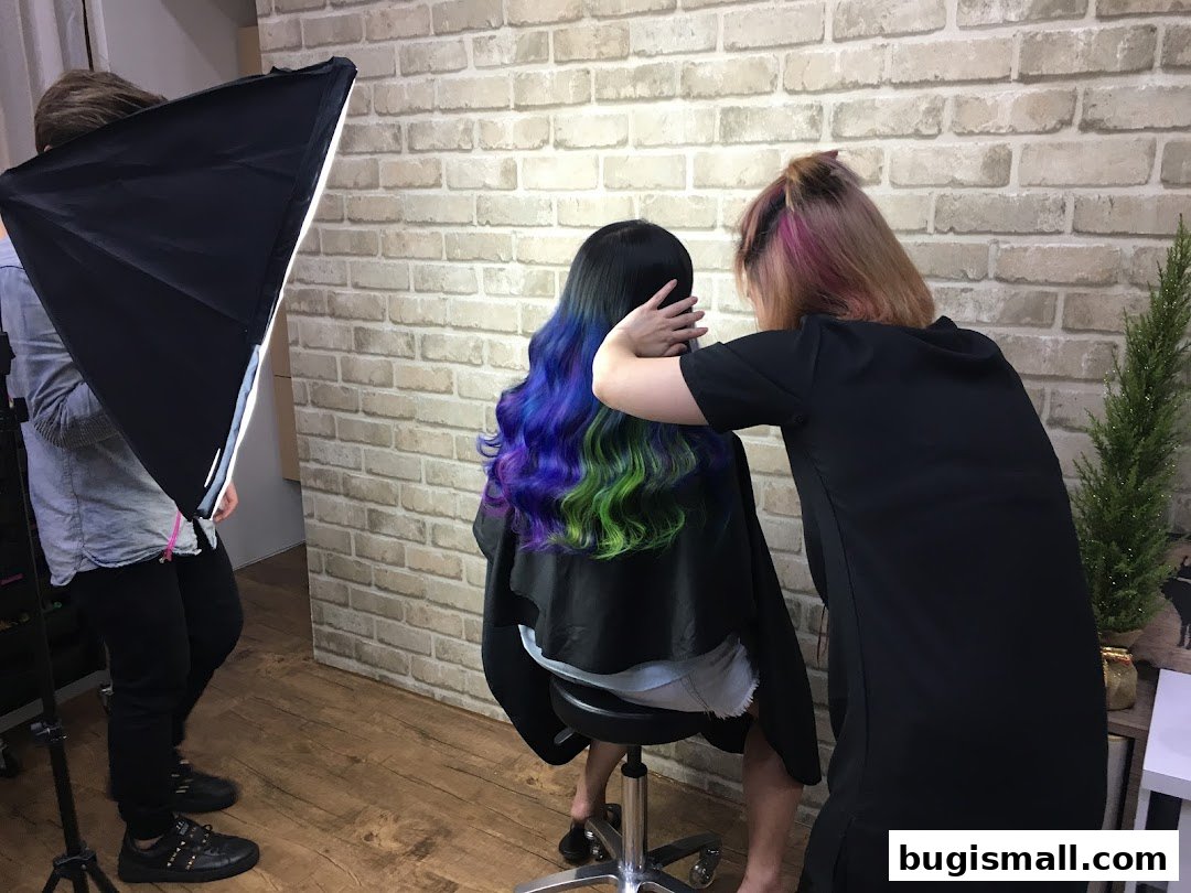 A hairstylist colors a client's hair with vibrant blue and green hues, while a photographer prepares lighting.