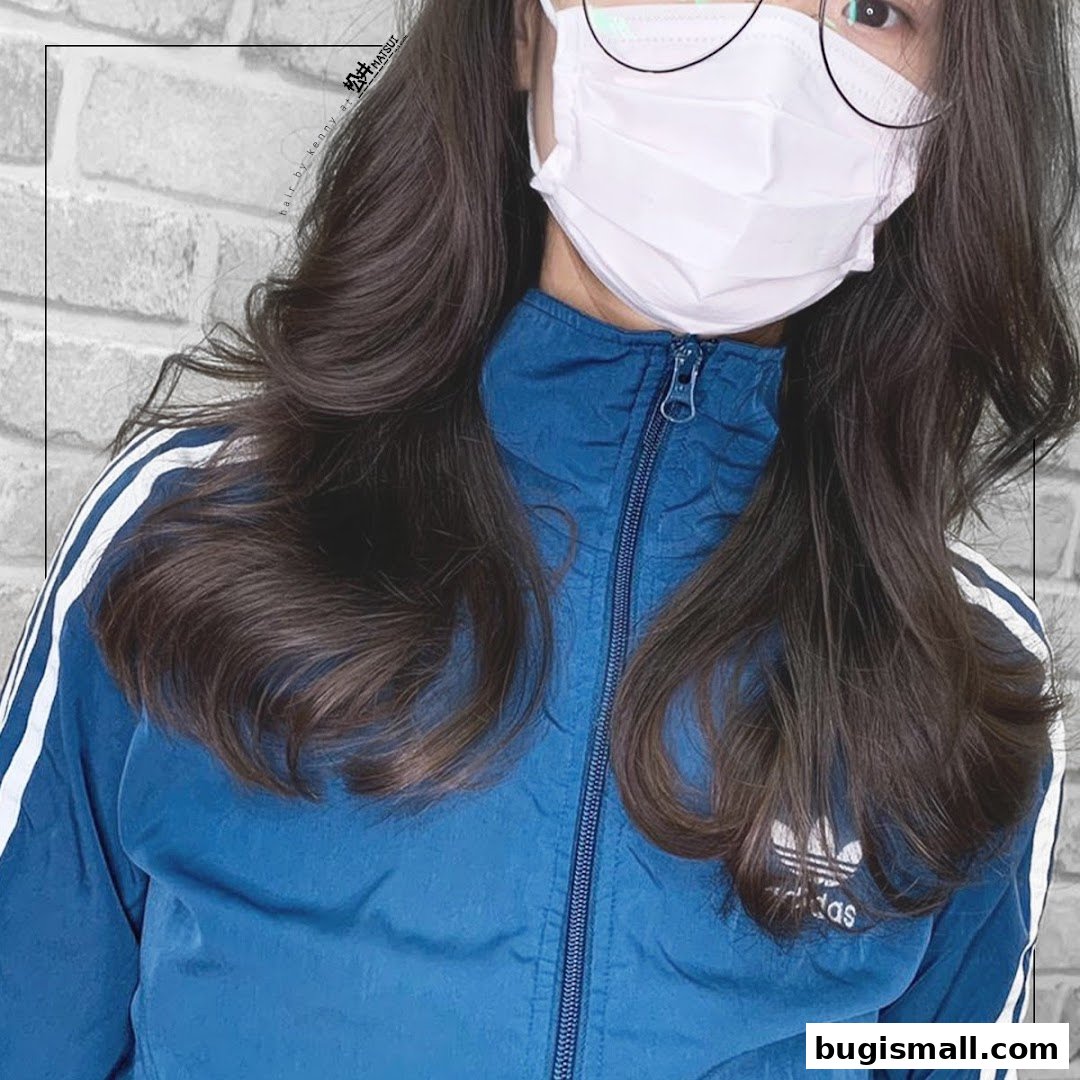 A person with long, dark, wavy hair wearing a blue jacket and a face mask.