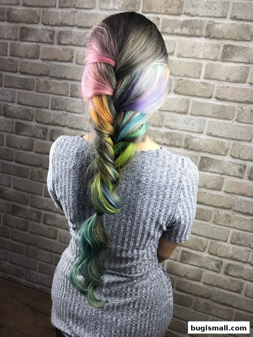 A person's back with a fishtail braid featuring rainbow hair colors against a brick wall.