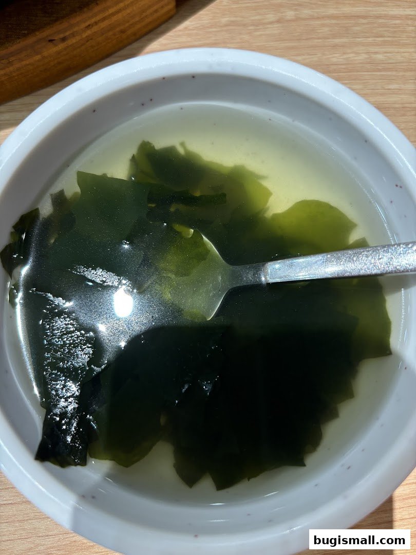 A close-up shot of a bowl of clear soup with large pieces of seaweed and a spoon.