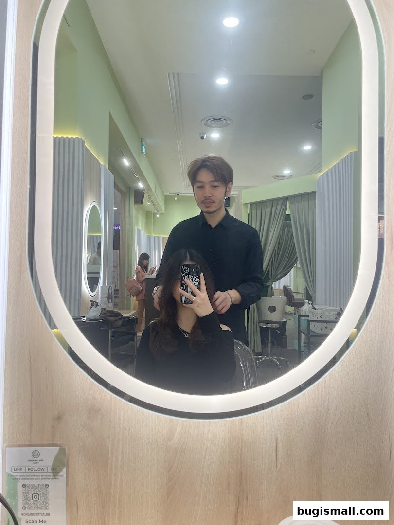 A hairstylist works on a client's hair in a modern salon, reflected in a large oval mirror.