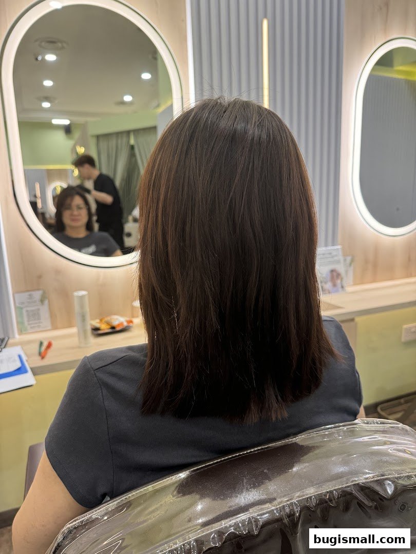 A person's back with dark hair sits in a salon chair, facing mirrors.