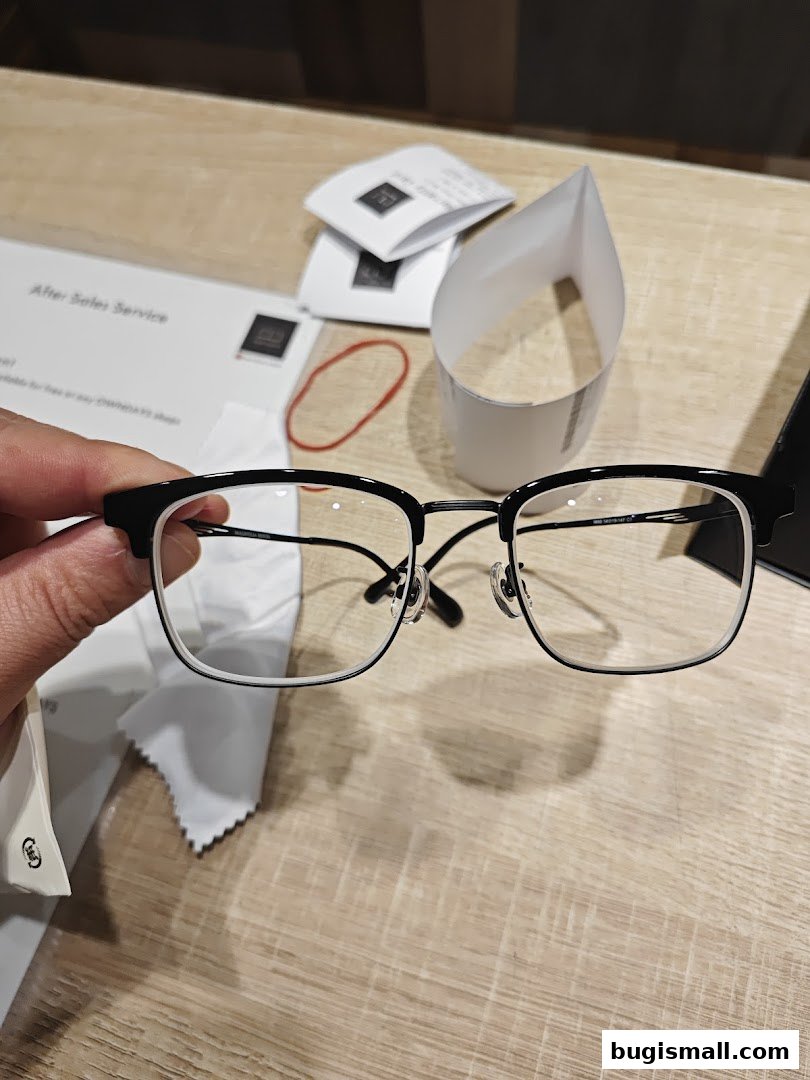 A hand holds a pair of black and white framed glasses with a cleaning cloth and papers on a wooden table.