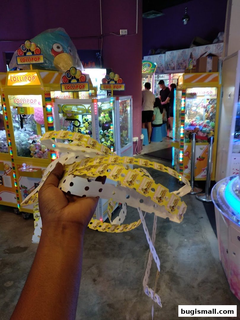 A hand holds a long strip of tickets in front of arcade claw machines.