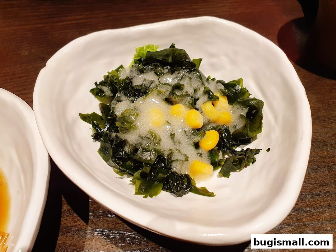 A close-up of a white bowl filled with seaweed salad and corn, topped with a foamy dressing.