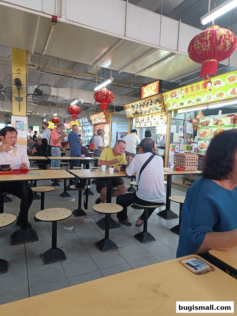 People dining at tables in a food court with Thai food stalls and red lanterns.