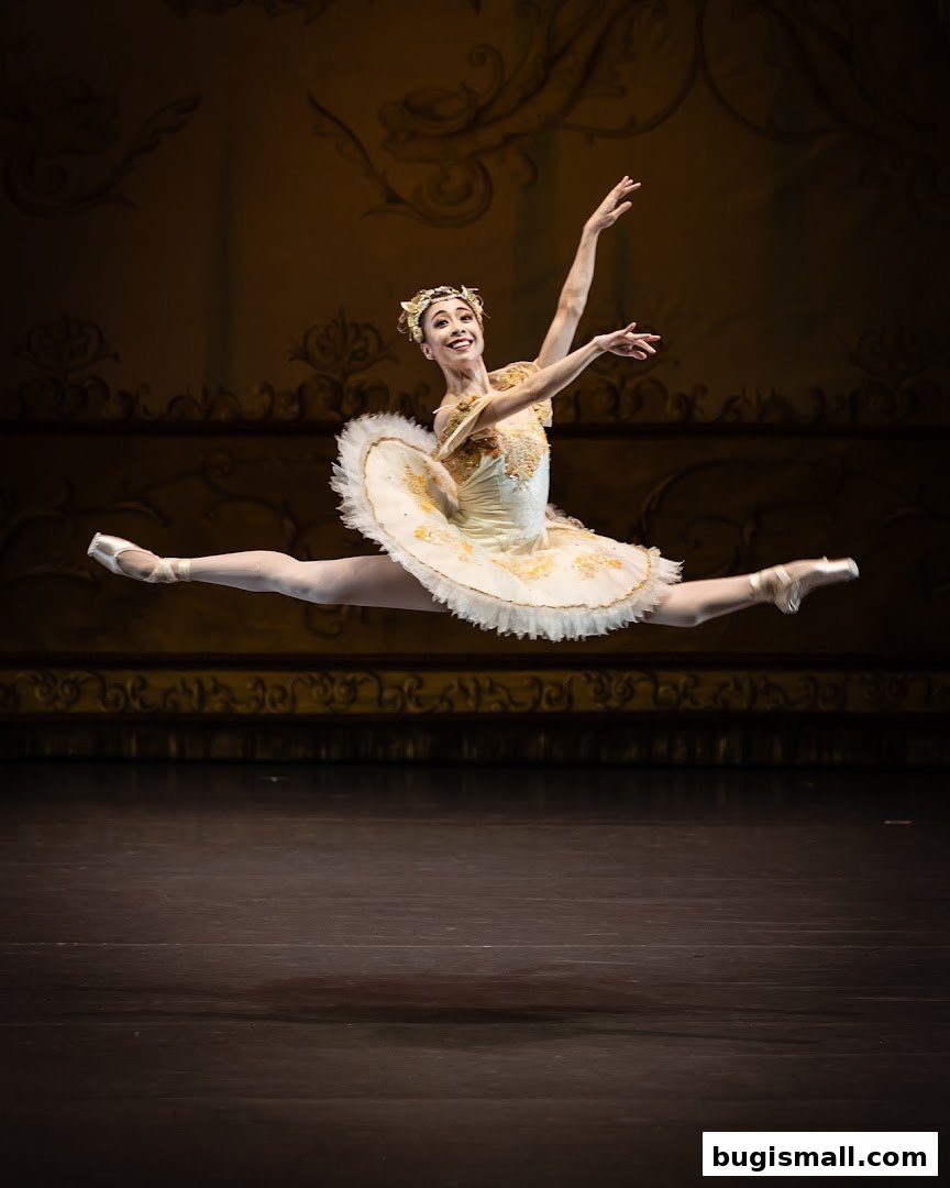 A ballerina in a white and gold tutu performs a split leap on a stage.