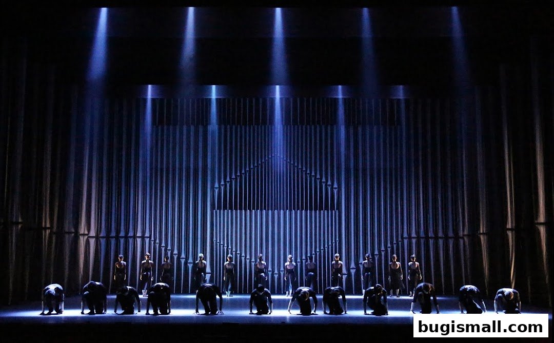 Dancers in black costumes perform on a stage with dramatic blue lighting and a pipe organ-like backdrop.