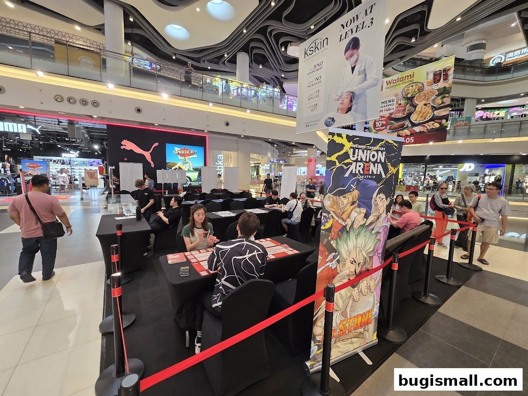 People playing a card game at tables in a mall with various advertisements overhead.