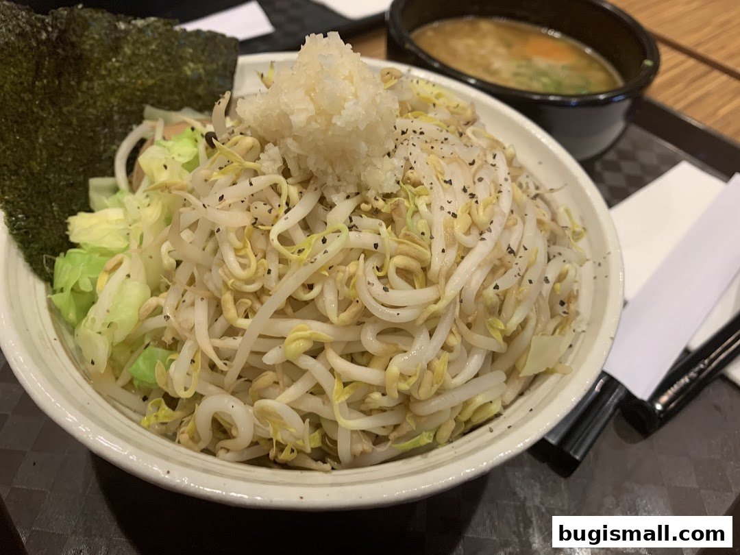 A bowl of ramen with bean sprouts, cabbage, nori, and minced garlic, served with a side of soup.