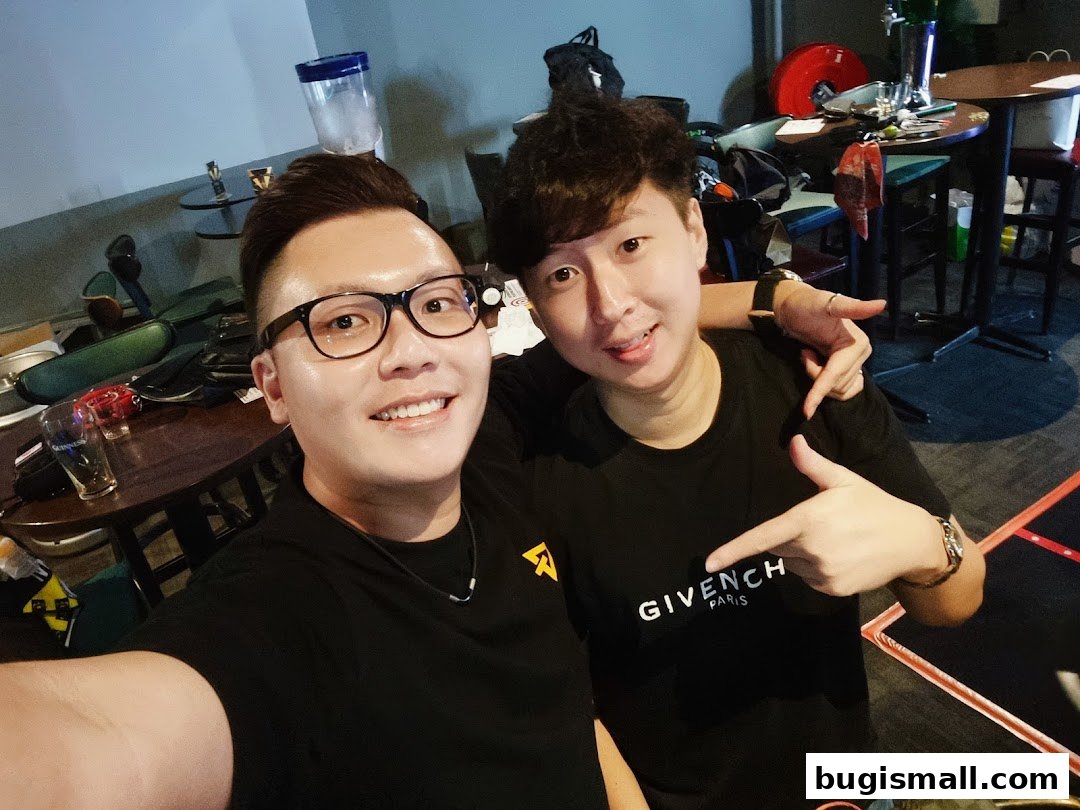 Two smiling men posing for a selfie in a bar with tables and chairs in the background.