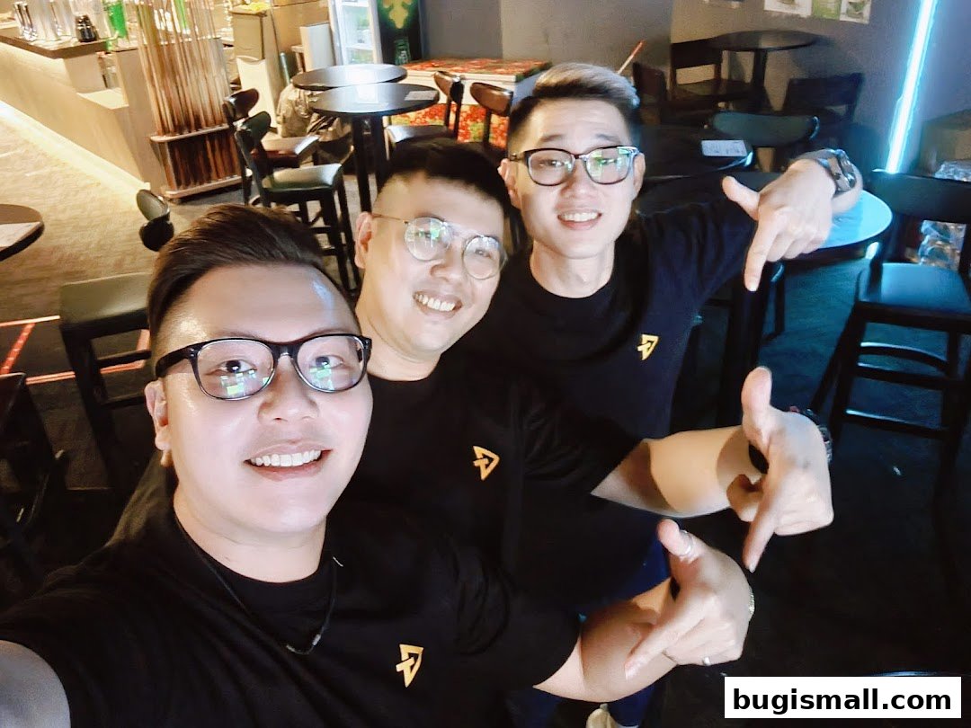Three smiling men in black t-shirts with a logo pose for a selfie in a dimly lit bar.