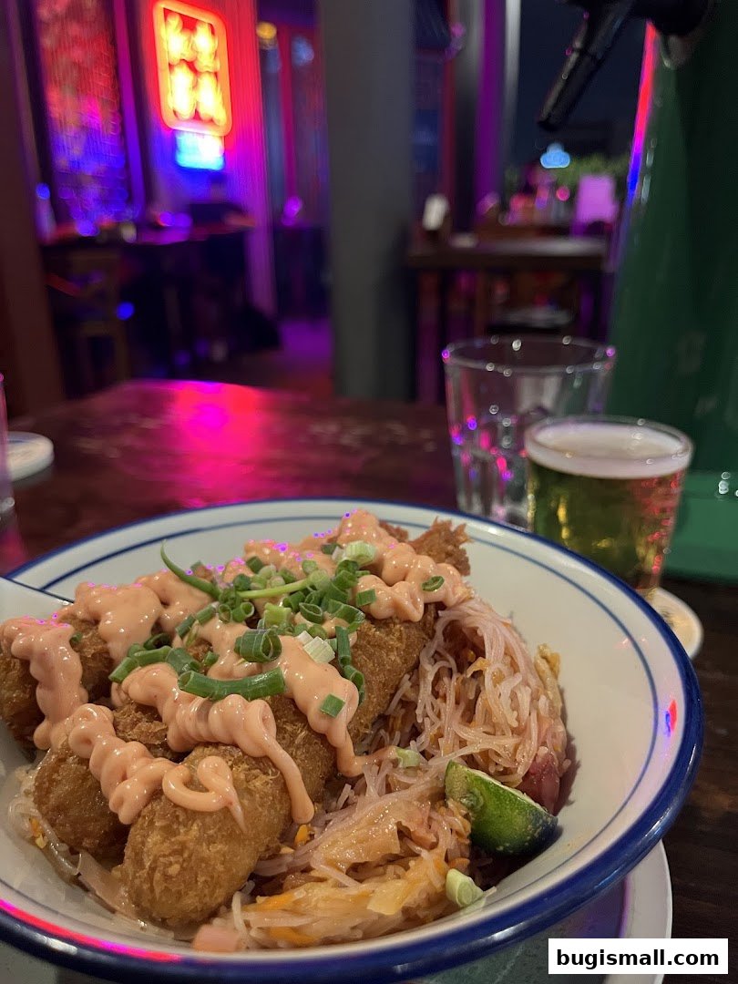 A bowl of food with fried items and noodles, served with a drink in a dimly lit bar.