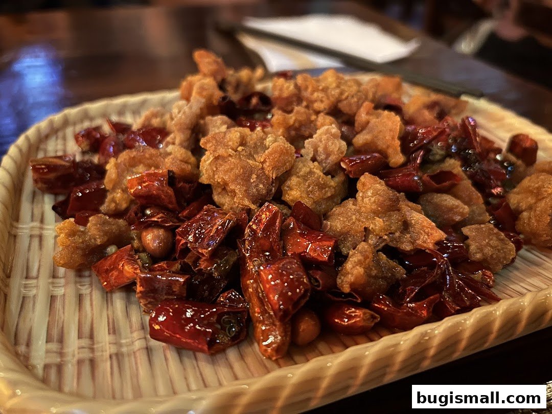 A close-up of crispy fried chicken bites with dried chili peppers and peanuts on a woven plate.