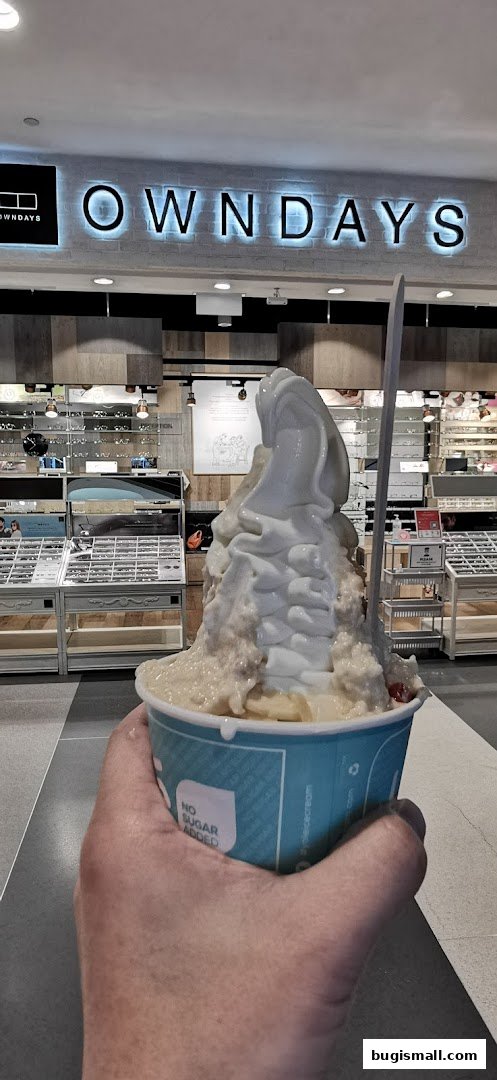 A hand holds a cup of soft-serve ice cream in front of an optical shop.