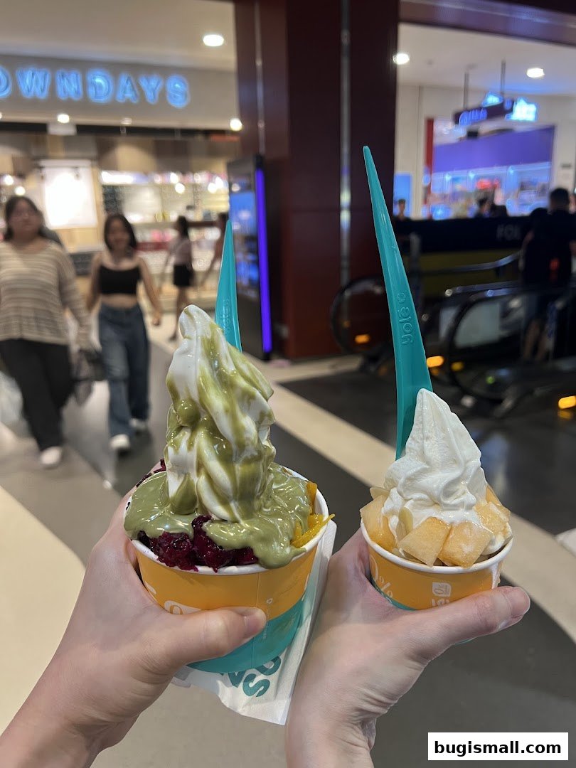 Two hands holding cups of froyo with toppings, with a mall interior in the background.