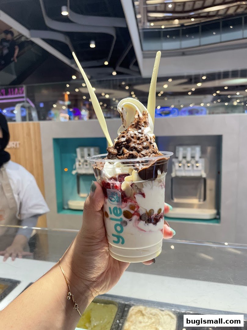 A hand holds a cup of frozen yogurt with toppings, including chocolate and fruit.
